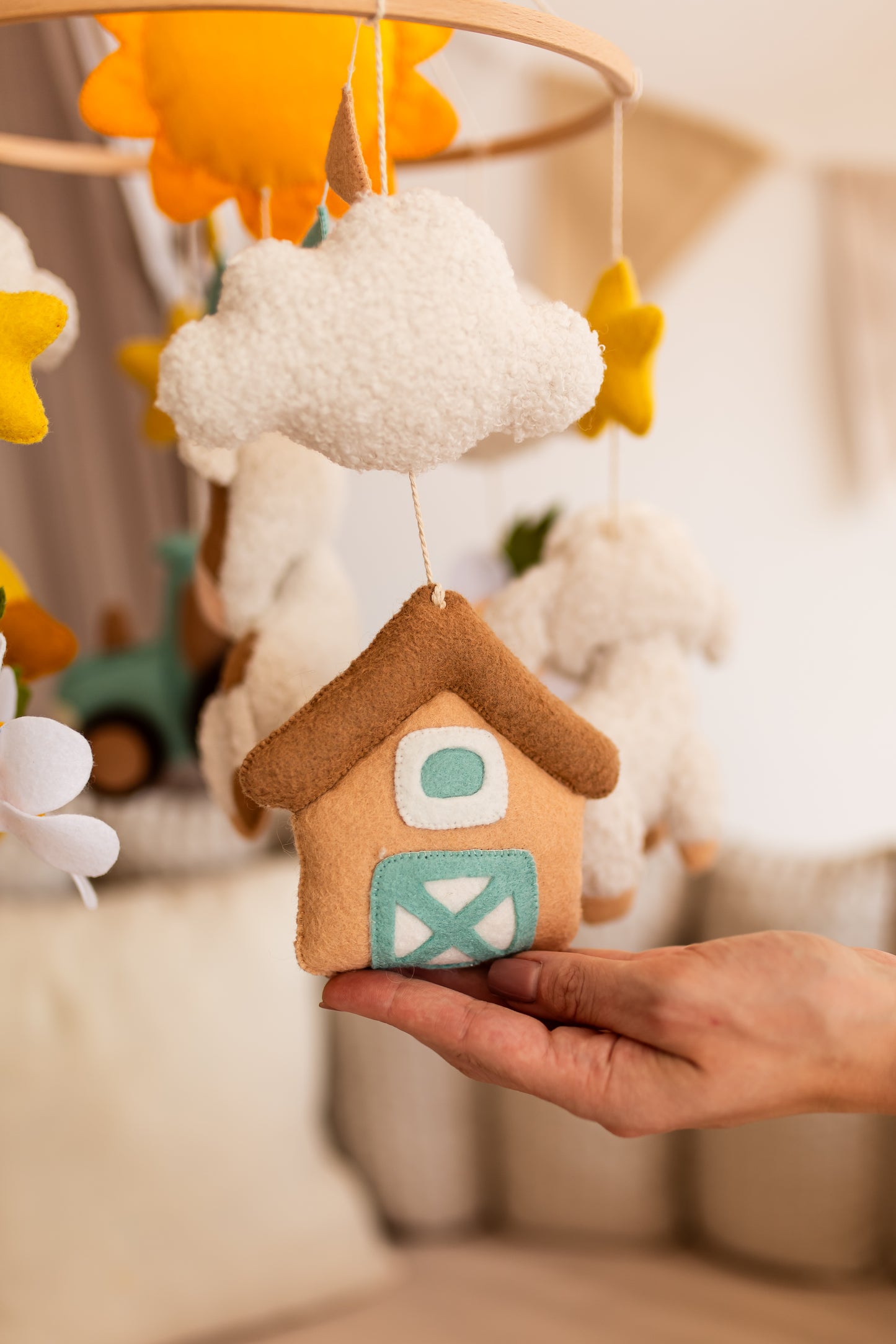 Hand holding a small house-shaped plush toy with a cloud and sun mobile in the background.