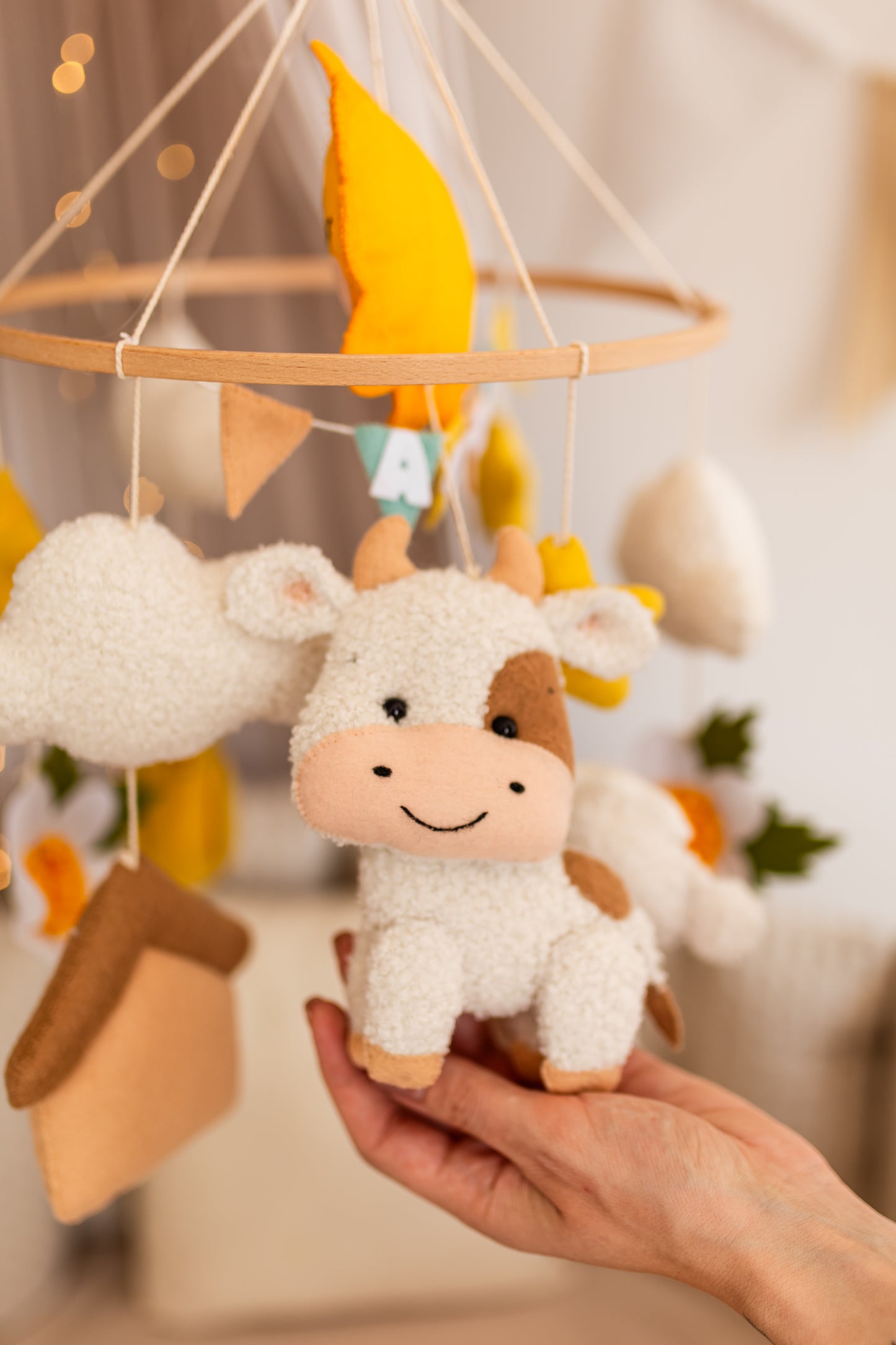 Plush cow toy held by a hand in front of a decorative mobile with yellow leaves and wooden elements.