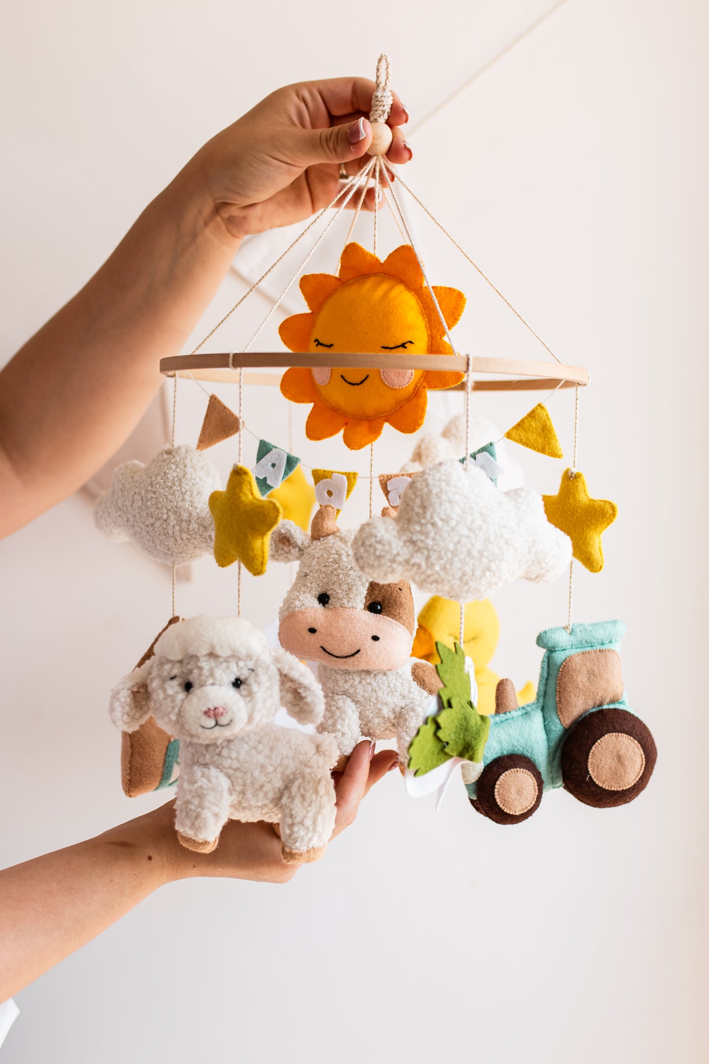 Plush toy mobile with sun, stars, sheep, and tractor held by a hand against a light background