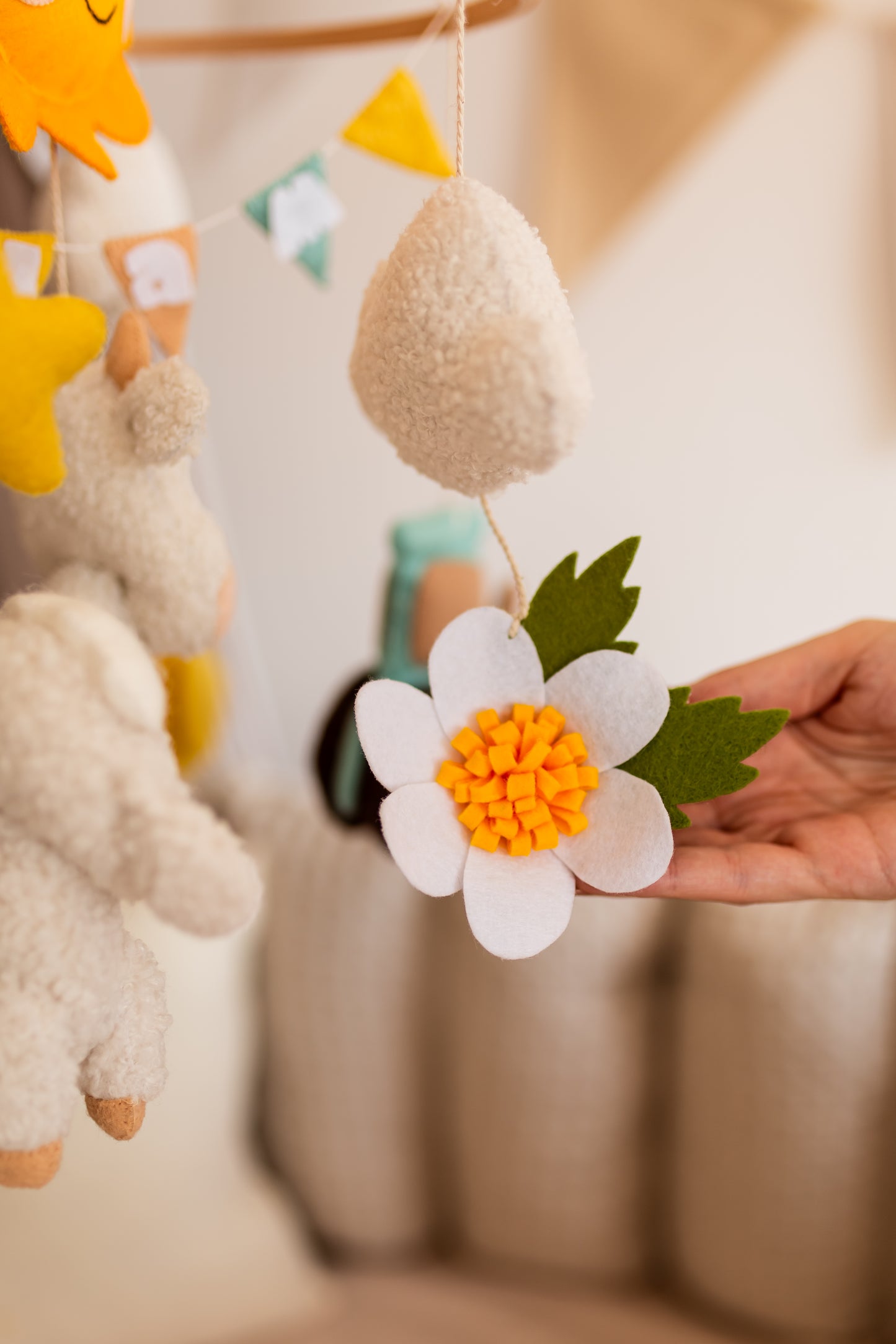 Close-up of a hand holding a decorative flower with a blurred background of a baby mobile.
