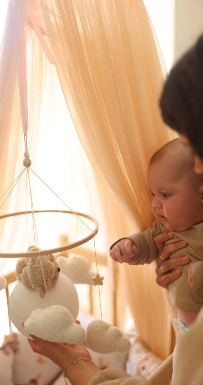 Baby held by an adult in a room with soft lighting, baby mobile and curtains.