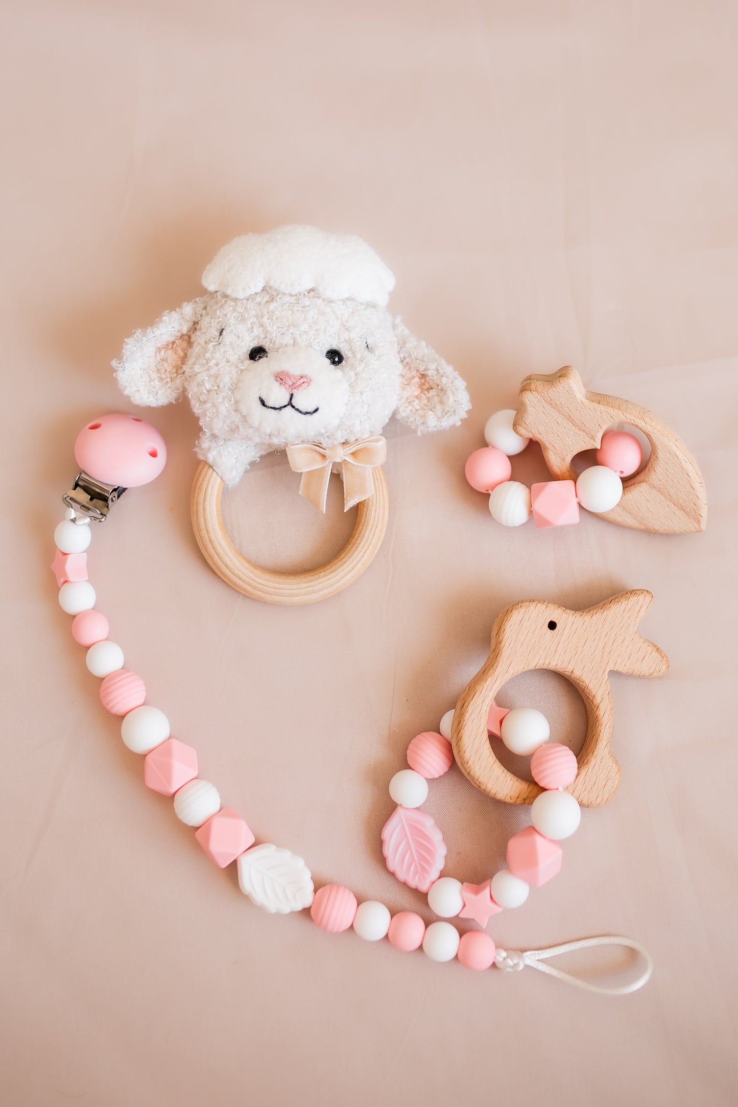 Baby teething toys including a lamb-shaped rattle, wooden teether, and pink bead necklace on a beige background.