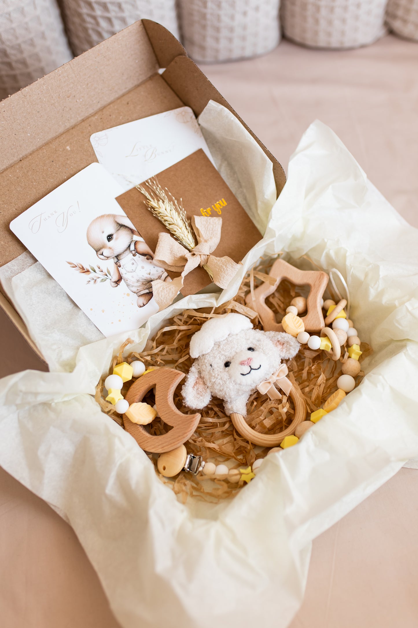 Gift box with baby-themed items including a plush toy, wooden stars, and cards on a neutral background.