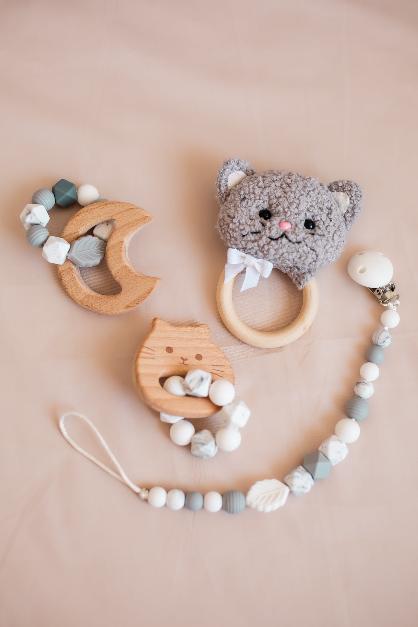 Set of baby teething toys including a wooden cat-shaped rattle, a gray cat-themed teether, and a necklace with beads on a beige background.